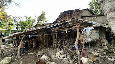 This is an image of a house severely damaged by rain. Two individuals are visible, one standing in the doorway and another just outside the structure, engaging in daily activities amidst these tough circumstances. 