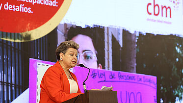 A woman, wearing a red jacket and short hair, is standing in front of a LED screen with CBM logo on it, and behind a podium, giving a speech. 