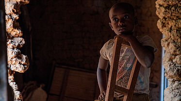 The picture shows a young girl standing in a dimly lit room with an expression of curiosity and contemplation. She is holding onto the back of a wooden walking stick. Sheeba is wearing a light-coloured T-shirt featuring a colourful design, and her short hair is neatly trimmed. Natural light coming through an opening highlight her face and upper body, creating a poignant and reflective atmosphere. Sheeba has a bilateral cataract and a physical impairment.