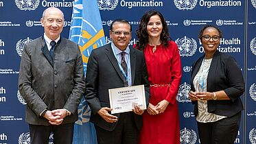 This image shows four individuals standing in front of a backdrop featuring the World Health Organization (WHO) logo. One of the individuals at the centre is holding a certificate that reads, "Certificate of Membership", awarded to the Christian Blind Mission (CBM). The people in the photo are dressed in formal attire and are smiling, indicating a celebratory moment for the recognition of CBM's membership to the WHO Global SPECS Network. This event highlights CBM's ongoing efforts and partnership in global health and disability inclusion.