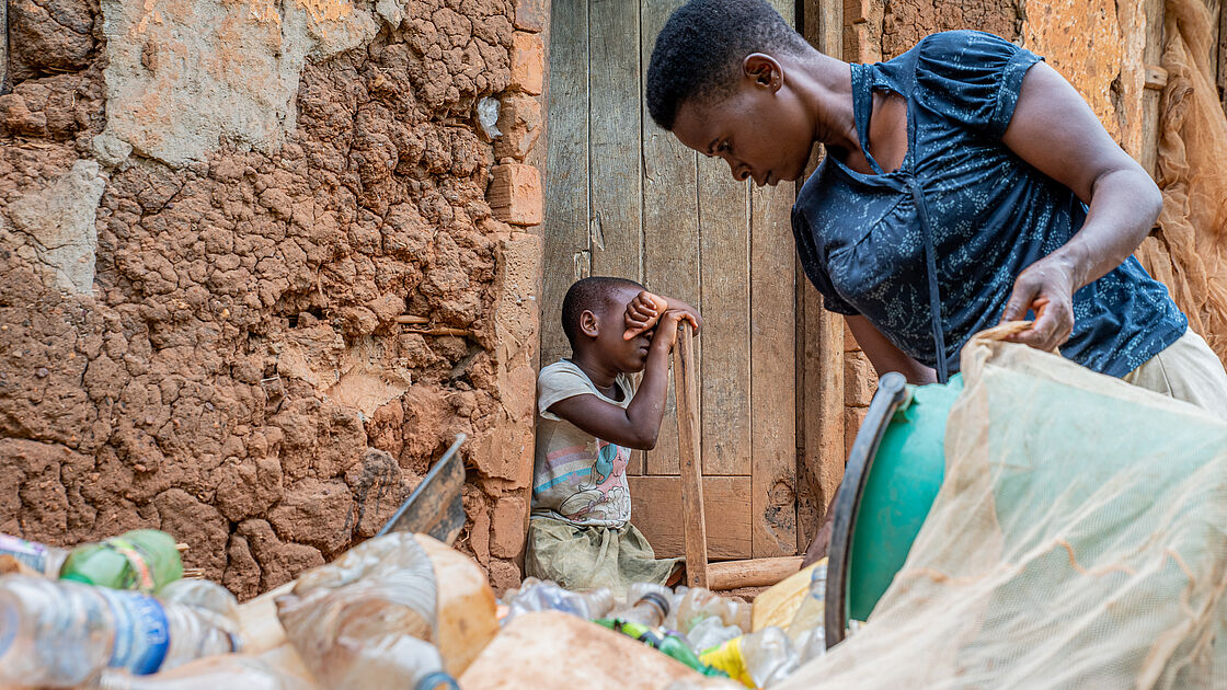 The picture shows a woman sorting through a pile of plastic bottles outside a rustic, clay-walled house. She is focused on her work. Nearby, a young girl leans against the doorway with one hand shielding her eyes from the bright sunlight. 